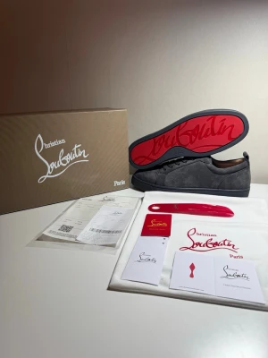 Gråa Christian Louboutin sneakers - Snygga gråa sneakers från Christian Louboutin i mocka med den ikoniska röda sulan och diskret logga på sidan. Skorna har lågt skaft och klassisk snörning. Perfekta för dig som vill sticka ut med exklusiv stil och detaljer.