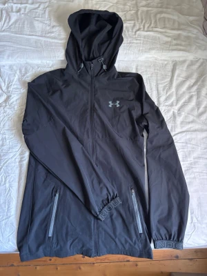 Underarmour vindbreaker - Svart vindjacka med huva från Under Armour. Jackan har hel dragkedja, två fickor med dragkedja och justerbar huva med snörning. Liten logga på bröstet och diskret tryck på ärmslutet. Perfekt för blåsiga dagar och sportiga outfits. Äkta och använd fåtal gånger utan täcken på andvändning