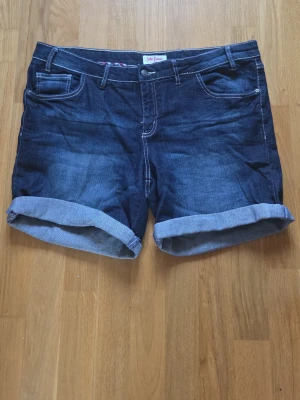 Jeansshorts från John Baner - Mörkblå jeansshorts från John Baner med klassisk femficksdesign och uppvikta benslut. Snygga kontrastsömmar och rutigt innerfoder i midjan ger en cool detalj. Perfekta för varma dagar och enkel att matcha med t-shirt eller linne.
