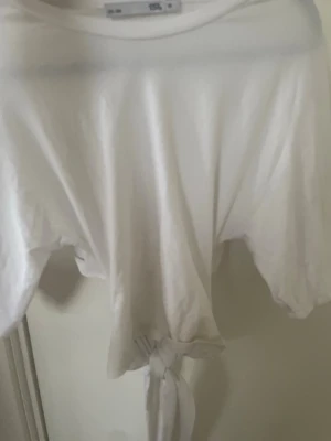 Vit croppad t-shirt med knyt - Säljer en vit croppad t-shirt med knyt framtill. T-shirten har korta ärmar och är tillverkad i mjuk bomull. Perfekt för en avslappnad och trendig stil.