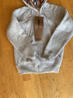 Grå hoodie från Burberry med dragkedja - Stilren grå hoodie från Burberry med hel dragkedja och klassisk snörning i huvan. Insidan av huvan har det ikoniska Burberry-mönstret. Perfekt för en avslappnad och trendig look.