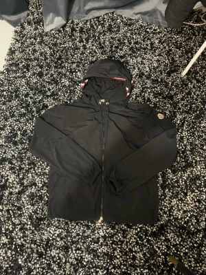 Svart vindjacka från Moncler - Snygg svart vindjacka från Moncler med huva och dragkedja framtill. Jackan har Moncler-logga på ärmen och detaljer i rött, vitt och blått på insidan av huvan. Tillverkad i lätt och slitstarkt material, perfekt för blåsiga dagar. Äkta!