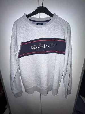 Grå sweatshirt från GANT - Snygg grå sweatshirt från GANT med klassisk logga framtill på en mörkblå panel med röda detaljer. Tröjan har rund halsringning, långa ärmar och ribbade muddar. Perfekt för en avslappnad och stilren look.