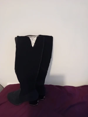 Svarta knähöga stövlar med klack - Snygga svarta knähöga stövlar i mockaimitation med bred klack och en cool V-formad detalj upptill. Perfekta för att lyfta vilken outfit som helst och ger en trendig siluett. Dragkedja på insidan för enkel påtagning.