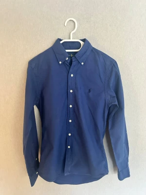 Blå skjorta från Ralph Lauren - Klassisk marinblå skjorta från Ralph Lauren med button-down krage och vita knappar. Skjortan har lång ärm och den ikoniska broderade loggan på bröstet. Perfekt för en stilren look och passar till många olika outfits. Använd fåtal gånger, schyssta priser vid snabb affär.
