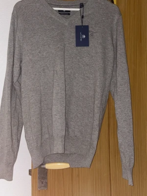 Grå V-ringad tröja från Gant - Snygg grå V-ringad tröja från Gant i premium bomull. Tröjan har långa ärmar, ribbade muddar och diskret broderad logga på bröstet. Perfekt för en stilren och avslappnad look.