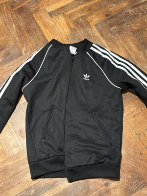 Svart Adidas vindjacka med vita ränder - Svart vindjacka från Adidas med klassiska vita ränder längs ärmarna och vit piping. Jackan har dragkedja framtill, två fickor med dragkedja och Adidas logga på bröstet. Sportig och stilren design, perfekt för en streetwear-look.