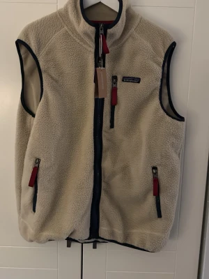 Beige fleeceväst från Patagonia - Snygg beige fleeceväst från Patagonia med marinblå detaljer och röda dragkedjor. Västen har hög krage, tre fickor med dragkedja och klassisk Patagonia-logga på bröstet. Perfekt för lager-på-lager och outdoor-stil.