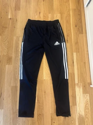 Svarta Adidas träningsbyxor - Säljer ett par svarta träningsbyxor från Adidas med klassiska vita ränder längs benen och Adidas-logga på låret. Byxorna har resår i midjan och dragkedjor vid bensluten. Perfekta för träning eller chill.