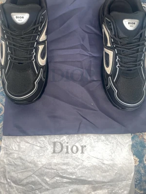Svarta Dior B30 sneakers - Säljer ett par svarta Dior B30 sneakers med vita och silverfärgade detaljer. Skorna har en chunky sula, mesh och läderpaneler samt tydlig Dior-logga på sidan. Snygg och modern design som sticker ut. Perfekta för dig som gillar exklusiva sneakers.