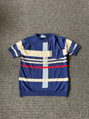 T-shirt - Snygg mörkblå t-shirt från Donville med beige, grå och röd randig design samt bröstficka. T-shirten har korta ärmar och är stickad i ett mjukt material som ger en vintagekänsla. Perfekt för dig som gillar retrostil och unika plagg.