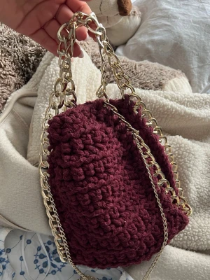 Vinröd virkad handväska med guldkedja - Trendig vinröd handväska i virkat garn med chunky guldfärgade kedjor som handtag och axelrem. Väskan har en mjuk, strukturerad form och ger en cool och unik look. Perfekt accessoar för att lyfta din outfit.