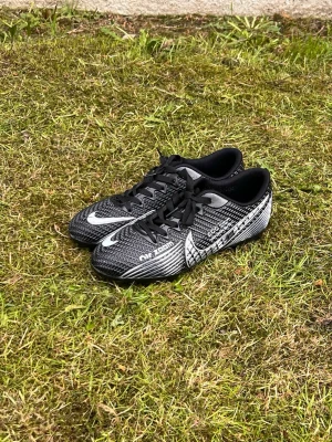 Nike Air Zoom svarta fotbollsskor - Snygga svarta Nike Air Zoom fotbollsskor med vita detaljer och klassisk snörning. Skorna har en texturerad ovandel för bättre bollkontroll och en låg profil som ger bra grepp på planen. Perfekta för dig som vill ha stil och prestanda på matchen.