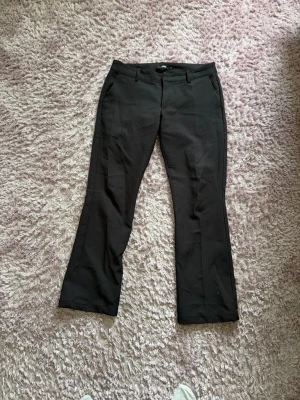 Svarta kostymbyxor med bootcut - Snygga svarta kostymbyxor med bootcut-modell och pressveck. Byxorna har hällor för skärp och sidofickor. Perfekta för en stilren look. Materialet ser ut att vara en klassisk kostymblandning. Lite noppriga