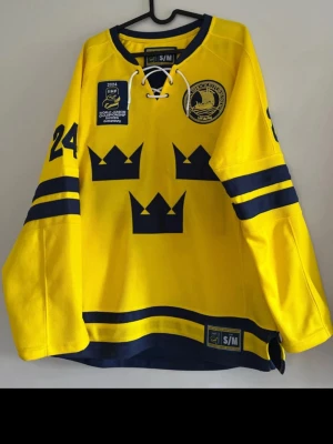 Gul Tre Kronor hockeytröja S/M - Gul hockeytröja från Svenska Ishockeyförbundet med Tre Kronor-tryck, blå detaljer och nummer 24 på ärmen. Tröjan har snörning i halsen, officiella IIHF World Junior Championship 2024-märke och är tillverkad i ventilerande syntetmaterial.
