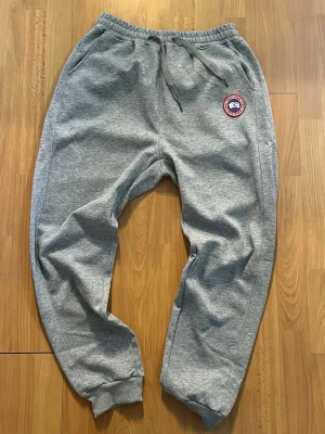 Grå mjukisbyxor från Canada Goose - Snygga grå mjukisbyxor från Canada Goose med broderad logotyp på vänster ben. Byxorna har dragsko i midjan och muddar vid bensluten. Tillverkade i mjukt bomullsmaterial för maximal komfort.