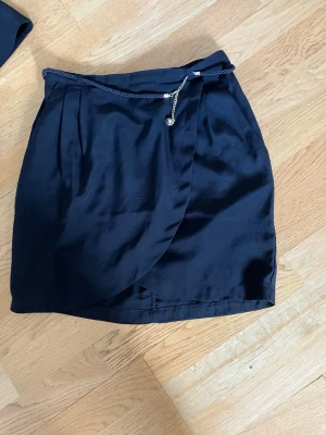 Svart omlottkjol med kedjedetalj - Snygg svart omlottkjol med dekorativ kedja i midjan. Kjolen har en kort passform och är perfekt för att skapa en trendig look. Materialet känns mjukt och följsamt, och kjolen har en stilren design med lätt glans.