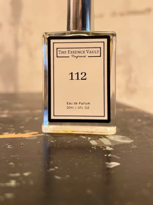 The Essence Vault 112 Eau de Parfum - Parfymflaska på 30 ml från The Essence Vault med doftnummer 112, inspirerad av Love, Don't Be Shy. Flaskan är rektangulär i genomskinligt glas med silverfärgad spraykork och innehåller en ljusgul vätska.
