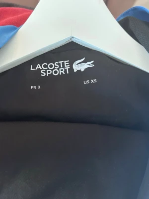 Svart sportjacka från Lacoste Sport XS-S - Snygg svart sportjacka från Lacoste Sport i storlek XS-S. Jackan har dragkedja och blå detaljer längs sidorna. Perfekt för träning eller sportiga aktiviteter. Klassisk design med Lacoste-logga på insidan.