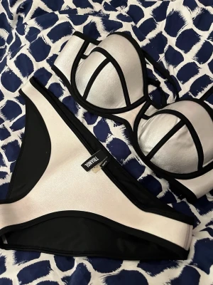 Silvrig bikini från Triangl - Snygg bikini från Triangl i metallic-silver med svarta kontrastkanter. Överdelen har bygel och vadderade kupor för extra stöd, och underdelen är klassisk brief-modell. Tillverkad i neoprenmaterial som ger en cool och sportig look. Den sitter som en större M/L, yttre tyget sitter inte fast i det tjockare neopren tyget på baksidan av nedredelen men det syns inte när den väl används och tyget stretchas.