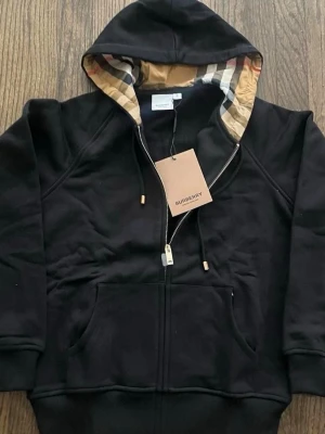 Svart hoodie från Burberry med dragkedja - Snygg svart hoodie från Burberry med klassiskt rutigt foder i huvan och dragkedja framtill. Storlek L men sitter som en M eller en större S. Hoodien har fickor på magen och ribbade muddar. Perfekt för dig som vill ha en stilren och exklusiv look med ikoniska Burberry-detaljer.