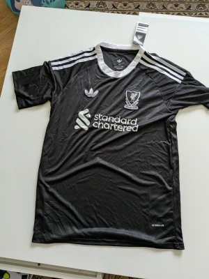 Svart Liverpool tröja från Adidas - Snygg svart Liverpool matchtröja från Adidas med vita detaljer och klassiska tre ränder på axlarna. Tröjan har klubbmärke, Adidas-logga och sponsortryck framtill. Tillverkad i lätt och ventilerande AEROREADY-material, perfekt för träning eller match.