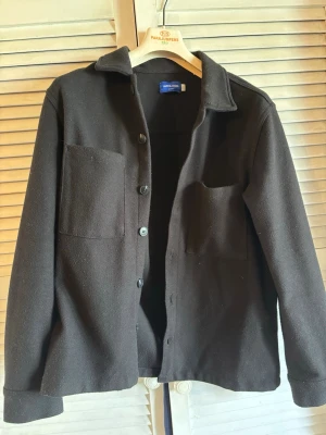 Svart overshirt från Jack & Jones - Stilren svart overshirt från Jack & Jones med klassisk krage, knäppning framtill och två stora bröstfickor. Overshirten har en rak passform och är tillverkad i ett mjukt tyg som passar perfekt för lager-på-lager-stil.