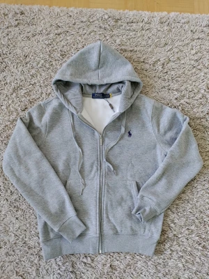 Grå hoodie från Polo Ralph Lauren - Snygg grå zip-hoodie från Polo Ralph Lauren med klassisk huva och dragsko. Broderad marinblå logga på bröstet och två fickor framtill. Tillverkad i mjuk bomullsblandning som känns skön mot huden. Perfekt för en chill och avslappnad stil.