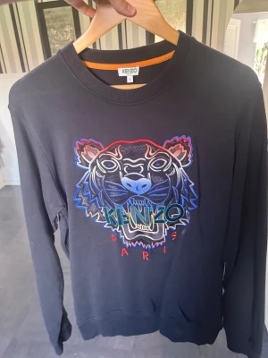 Svart Kenzo sweatshirt med tiger - Svart sweatshirt från Kenzo med ikoniskt broderat tigerhuvud i blått, rött och vitt framtill. Rund halsringning, ribbade muddar och klassisk passform. Perfekt statement-plagg för dig som gillar streetwear och snygga detaljer.