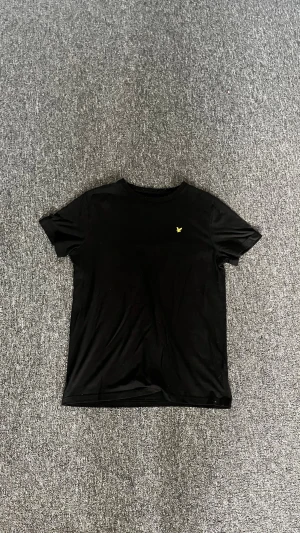 Svart t-shirt från Lyle & Scott - Klassisk svart t-shirt från Lyle & Scott med gul broderad logga på bröstet. T-shirten har rund halsringning och korta ärmar. Tillverkad i mjuk bomull för en skön känsla och enkel stil.