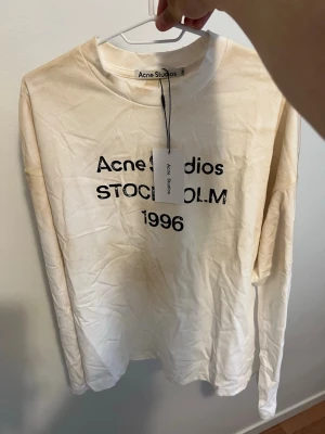 Vit långärmad t-shirt Acne Studios - Vit långärmad t-shirt från Acne Studios med tryck i svart text på bröstet där det står 'Acne Studios STOCKHOLM 1996'. Klassisk rund halsringning och avslappnad passform. Tillverkad i mjuk bomull och har en enkel, stilren design.