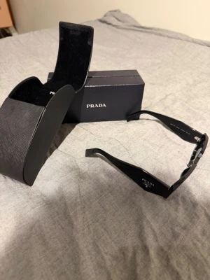 Svarta solglasögon från Prada - Snygga svarta solglasögon från Prada med rektangulära bågar och breda skalmar. Kommer med originalfodral och ask. Perfekta för dig som vill ha en stilren och lyxig accessoar till din outfit.