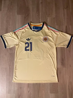 Chong Curacao VM-2026 Fotbollströja - Snygg gul Curacao VM-2026 Fotbollströja med nummer 21 och namnet Chong på ryggen. Tröjan har färgglada ränder på axlarna, mörkblå detaljer vid ärmslut och krage samt klubbmärke på bröstet. Perfekt för dig som gillar sportig stil. HELT NY!