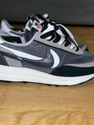 Nike x Sacai LDWaffle svart/grå DISCONTINUED  - Nike x Sacai LDWaffle sneakers i svart, grå och vit. Skorna har dubbla Swoosh-loggor, mesh- och mockadetaljer samt dubbla skosnören och tungor. Snygg retrostil med låg profil och chunky sula. Perfekta för dig som gillar streetwear och unika detaljer.