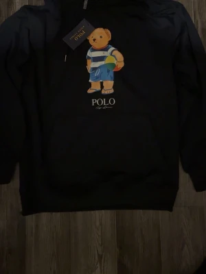 Svart hoodie Polo Ralph Lauren björn - Svart hoodie från Polo Ralph Lauren med den ikoniska björnen på bröstet, klädd i blåvitrandig tröja och blå shorts. Hoodien har huva med dragsko och stor magficka framtill. Mjuk bomullskvalitet och klassisk passform. Perfekt för en chill och stilren look.