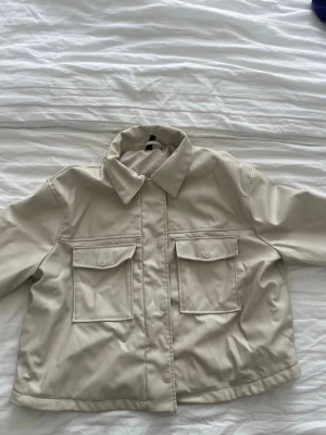 Beige läderjacka - Stilren beige läderjacka med två stora bröstfickor och knappar framtill. Den har klassisk krage och raka ärmar. 