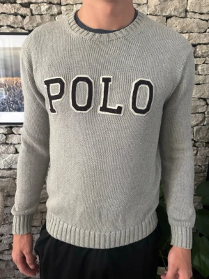 Ralph Lauren stickad  - Säljer en grå stickad tröja från Polo med stora svarta bokstäver på bröstet. Modellen är 188 och väger 90kg, har oftas L i kläder! 