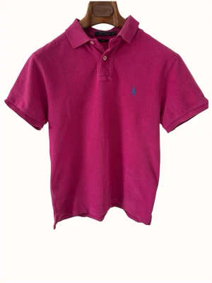 Rosa pikétröja från Ralph Lauren - Snygg rosa pikétröja från Polo Ralph Lauren i slim fit-modell. Tröjan har korta ärmar, klassisk krage och två knappar framtill. Den ikoniska blå loggan är broderad på bröstet. Perfekt för en avslappnad men stilren look.