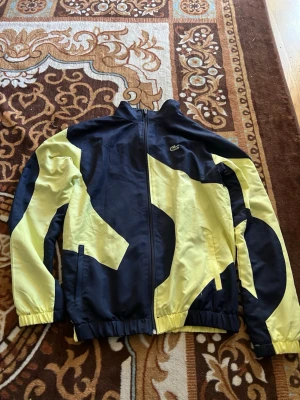 Svart och gul vindjacka från Lacoste - Snygg vindjacka från Lacoste i svart och gul med coolt grafiskt mönster. Jackan har hel dragkedja, hög krage och elastiska muddar vid ärmar och midja. Perfekt för dig som vill sticka ut med en sportig och trendig look.
