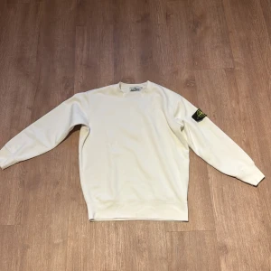 Vit sweatshirt från Stone Island - Vit hoodie från Stone Island med klassisk rund hals och långärmad passform. Tröjan har den ikoniska Stone Island-loggan på vänster ärm, vilket ger en snygg streetwear-vibe. Tillverkad i mjuk bomull för skön komfort och stil.