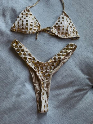 Originell bikini med guldprickar, helt oanvänd. - Supersnygg och originell bikini med beige/hudfärgad botten med glansiga guldprickar. Klassisk trekantsmodell upptill med knyt i nacken och ryggen, samt matchande bikiniunderdel med högt v-skuren passform. Perfekt för strandhäng och poolparty! Aldrig använd. Ärliga och snabba affärer uppskattas:)