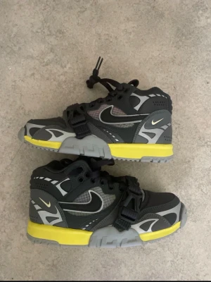 Nike svarta och grå sneakers med gult - Snygga sneakers från Nike i svart och grått med detaljer i gult och vitt. Skorna har en robust design med meshpaneler, kardborrespänne och klassisk Swoosh-logga på sidan. Sulan är grå med en gul rand som sticker ut. Perfekta för dig som gillar streetwear och sportig stil.