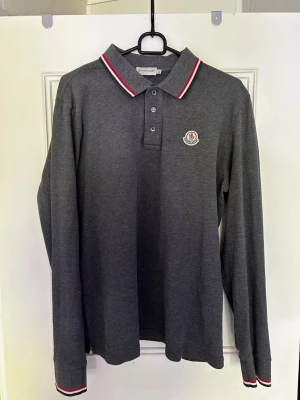 Mörkgrå långärmad polotopp Moncler - Mörkgrå långärmad pikétröja från Moncler. Tröjan är i ett bra skick och har används bara några gånger. Köptes från Merchsweden förra året. Tröjan är i storleken XL men passar L. 
