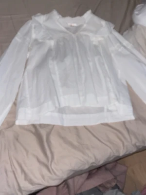 Vit blus med volangkrage - Söt vit blus med volangdetaljer vid kragen och v-ringning. Blusen har långa ärmar och är gjord i ett tunt, lätt material som ger en luftig känsla. Perfekt att styla med jeans eller kjol för en trendig look.