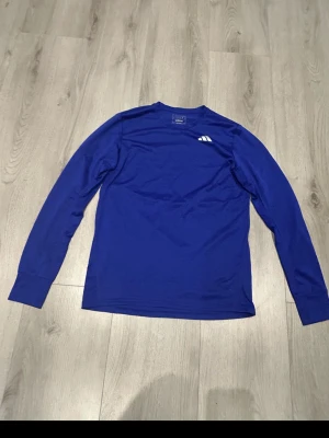 Blå långärmad träningströja Adidas - Snygg blå långärmad träningströja från Adidas med vita detaljer, inklusive tre ränder på ryggen och liten logga på bröstet. Tillverkad i ett lätt och funktionellt material som passar perfekt för träning och sportaktiviteter.