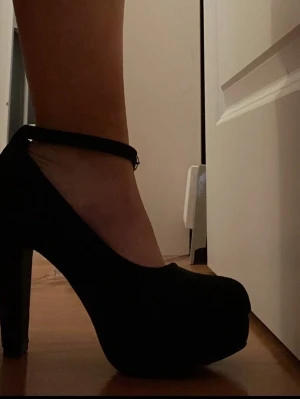 Svarta pumps med ankelrem - Snygga svarta pumps med hög klack och platåsula. Skorna har rundad tå och justerbar ankelrem med spänne. Materialet ser ut att vara mockaimitation som ger en matt och stilren look. Perfekta för att lyfta vilken outfit som helst. Otroligt sköna för att vara klackskor men tyvärr för stora för mig då jag har storlek37.