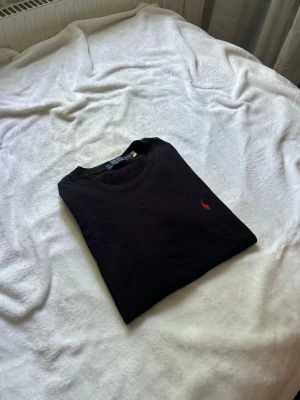 Svart t-shirt från Polo Ralph Lauren - Svart t-shirt från Polo Ralph Lauren med klassisk rund hals och liten röd broderad logga på bröstet. Enkel och stilren design som passar till det mesta. Tillverkad i mjuk bomull för en skön känsla.
