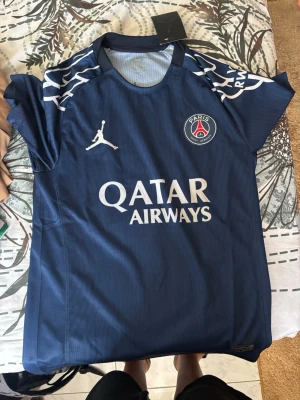 PSG matchtröja blå Jordan - Snygg blå PSG matchtröja från Jordan med vita detaljer och logga på bröstet. Tröjan har Qatar Airways tryckt på framsidan och coola vita mönster på axlarna. Perfekt för dig som älskar fotboll och vill representera Paris Saint-Germain.