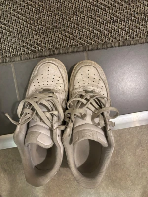 Vita Nike Air Force 1 sneakers - Klassiska vita Nike Air Force 1 sneakers med perforerad tåbox och högt skaft. Skorna har en chunky sula och är tillverkade i läder med textildetaljer. Perfekta för dig som gillar streetwear och tidlös stil.