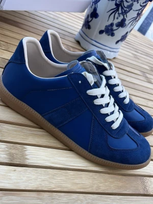 Maison Margiela  - Snygga Maison Margielas med vita skosnören och beige sula. Skorna har en mix av mocka och slätt material, samt diskreta paneler på sidorna. Insidan är ljus och de har en klassisk låg profil som passar till många olika stilar. (Vet ej äktighet)